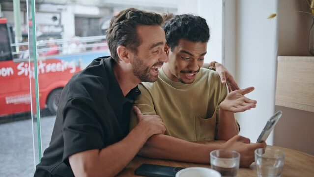 Joyful gays video calling looking smartphone at cafeteria closeup. Lgbt couple