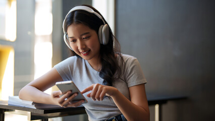 Asian teenage student woman wearing headphone to watching movie and surfing social media on smartphone to relaxation after studying and learning education knowledge for preparing test exam in cafe