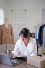 Young asian merchant women checking purchase online order and client information on laptop to taking notes in clipboard while working and shipping delivery for online shopping business in home office - 788990848