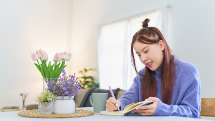 Asian business woman in sweater analyzing about strategy of new business and writing business project plan in notebook during planning about finance and market of new startup while working from home