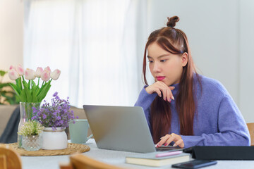 Asian business woman in sweater reading data on laptop to analyzing marketing strategy of project and typing business report during working about business plan of new startup while working from home
