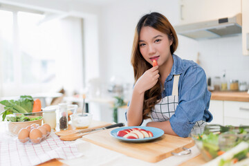Young asian woman enjoying and eating slices apple while preparing fruits and fresh vegetables salad to cooking healthy breakfast food for homemade snack in kitchen with healthy lifestyle at home