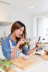 Young asian woman eating yogurt and use smartphone to searching food recipes while preparing fresh vegetables salad and cooking healthy breakfast food in modern kitchen with healthy lifestyle at home