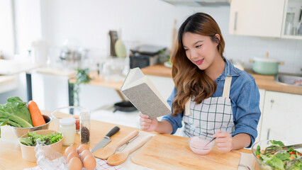 Young asian woman eating delicious yogurt and reading food recipes on a book while preparing fresh vegetables salad and cooking healthy breakfast food in modern kitchen with healthy lifestyle at home - 788991265