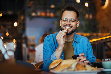 This is so delicious. Young man sitting in a cafe and enjoying in breakfast. Food, lifestyle concept. Happy young man having hamburger and french fries in fast food restaurant. - 788997678