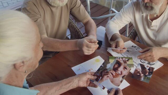 High-angle footage of three retirement home residents sitting around table, senior lady showing photos of grandkids and family to male companions, admiring and talking about them