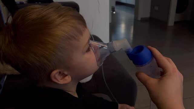Close up of little caucasian sick boy holding a inhalation mask on his face to breath saline solution, at home