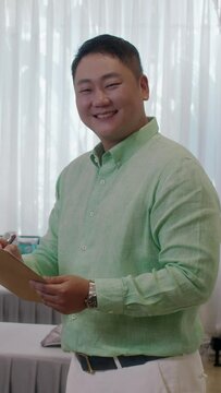 Vertical portrait of positive male Asian beauty salon manager with document folder smiling at camera