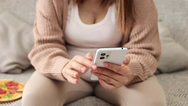Unrecognizable expecting lady wearing casual clothing sitting on sofa using cell phone touching gadget screen shopping on Internet using mobile app for pregnant women