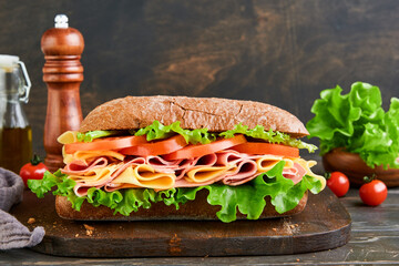 Sandwich. One fresh big submarine sandwich with ham, cheese, lettuce, tomatoes and microgreens on old wooden dark background. Healthy breakfast theme concept, school lunch, breakfast or snack.
