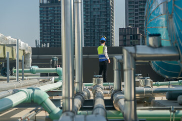 Focused engineer in hard hat and reflective vest standing amidst rooftop mechanical structures,...