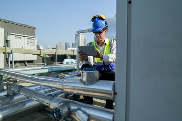 Young technician with hard hat and safety vest adjusts equipment on a commercial building's rooftop amidst an urban landscape under a clear sky.