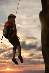 A woman is climbing a rock wall with a rope. The sky is cloudy and the sun is setting