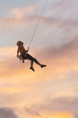 A woman is hanging from a rope, and the sky is orange and pink. Scene is adventurous and exciting