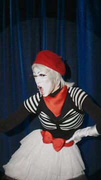 Vertical shot of funny female mime artist imitating weightlifting exercises with flower toy as dumbbell and showing biceps on camera during comedy performance on stage