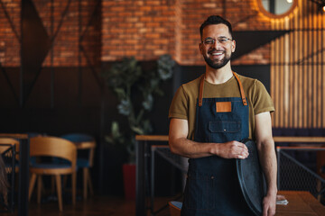 Portrait of Owner / Waiter at Restaurant. Portrait of handsome young male coffee shop owner standing at cafe. Portrait of a handsome barista in t-shirt and apron - 789034828