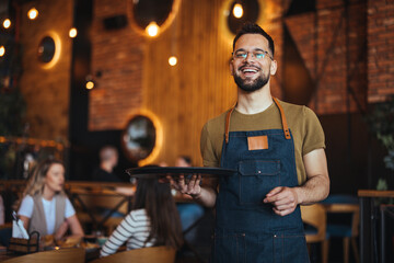 Waist up portrait of handsome waiter smiling cheerfully at camera standing in restaurant or cafe,...