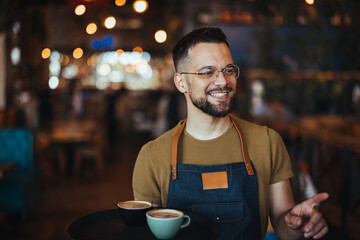 Waiter carrying tray with cappuccino cups in a cafe. Close up of handsome barista holding two cups of coffee in the cafe. Young waiter serving coffee in a cafe and looking at camera. - 789036074