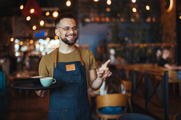 Portrait of smiling young waiter holding tray in cafe. Handsome waiter smiling at camera holding tray at the coffee shop. Profesional Waiter in Restaurant - 789036212