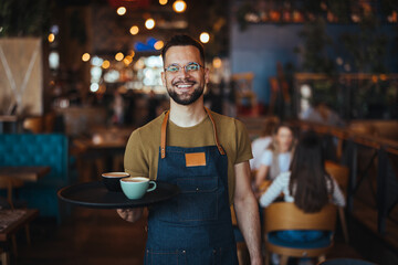Happy waiter holding a tray while working in cafe and looking at camera. Young waiter serving coffee in a cafe and looking at camera. - 789036237