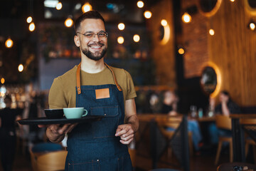 Portrait of Owner / Waiter at Restaurant. Portrait of handsome young male coffee shop owner standing at cafe. Portrait of a handsome barista in t-shirt and apron - 789036471