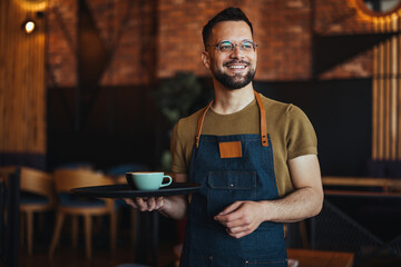 Happy waiter holding a tray while working in cafe and looking at camera. Young waiter serving coffee in a cafe and looking at camera. - 789036861