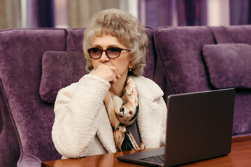 An elderly woman works on a laptop while sitting at a table in a cafe on a purple background