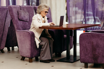 An elderly woman works on a laptop while sitting at a table in a cafe on a purple background