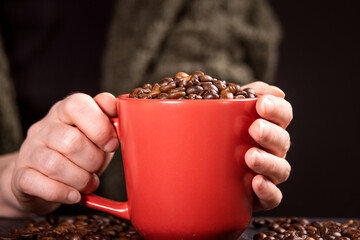 a lot of macro aromatic coffee beans in a red cup in female hands on a dark background.