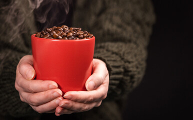 a lot of macro aromatic coffee beans in a red cup in female hands on a dark background.