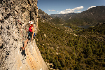 A woman is climbing a rock wall with a red shirt and a yellow helmet - obrazy, fototapety, plakaty