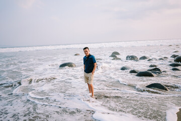 A contented man in a blue tee and shorts stands by the shore, his gaze fixed towards the horizon, radiating happiness against the backdrop of the beach