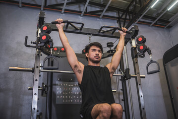 A strong and fit Asian man engages in hanging knee raises at a gym, targeting his core and abs for...