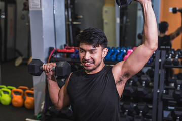 An asian man in a black tank top does seated alternating dumbbell presses. Training shoulders at the gym.