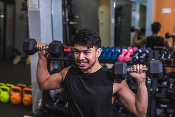 An asian man in a black tank top does seated dumbbell presses. Training shoulders at the gym.