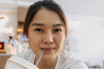 Close up of asian Thai woman smiling and taking selfie at cafe while drinking coffee beverage from straw.