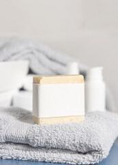 Soap bar with blank label on light grey towel near basin and cream bottles in bath, mockup
