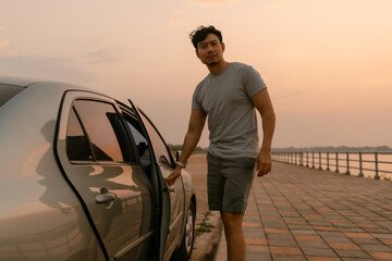 Asian man parking a car near riverside after long drive, looking at camera while closing car door...