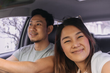 Front view of asian Thai couple, lover traveling by a car, woman taking selfie while man is driving on road. Family vacation concept.