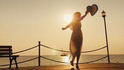 A woman meets the dawn at sea. Emotionally spinning with a hat in his hand on the pier. Dream of travel concept