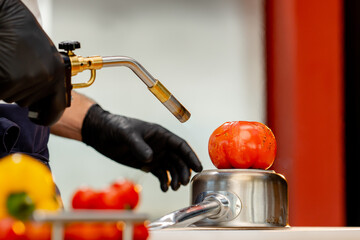 close-up, the chef stands in the kitchen wearing black gloves holds burner and burns a tomato