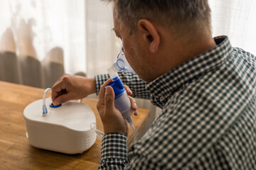 Elderly man sitting on a table and using a nebulizer mist at home - obrazy, fototapety, plakaty