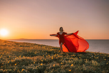 woman red dress is standing on a grassy hill overlooking the ocean. The sky is a beautiful mix of...