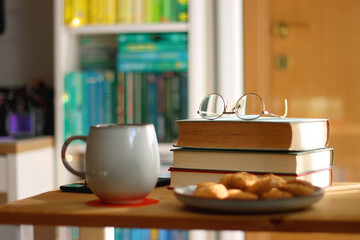 Cup of tea or coffee, pile of books, plate of cookies, reading glasses, e-reader and pen on the table. Colorful rainbow bookshelf in the background. Selective focus.