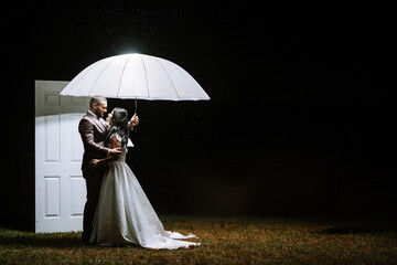 Valmiera, Latvia- July 28, 2023 - A bride and groom stand close under a white umbrella at night...
