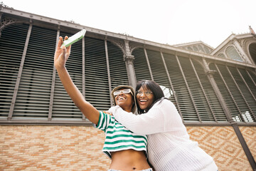 Portrait of two young cheerful black women embracing and taking a selife in an outdoor setting.