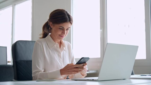 Smiling mature business woman executive using mobile phone working in office. Happy middle aged businesswoman holding smartphone, professional female executive looking at cellphone at workplace.