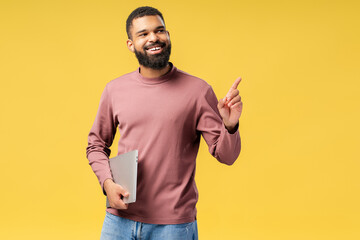 Happy young African American man pointing by finger while holding laptop, over yellow background