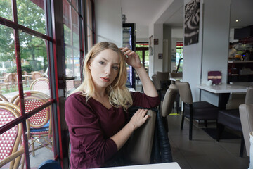 junge blonde frau sitzt einsam und allein an einem fenster in eiem cafe - obrazy, fototapety, plakaty