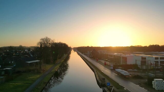 This drone footage captures the first light of daybreak reflecting on a calm canal, flanked by a road busy with early morning commuters. The rising sun casts a golden glow over the scene, contrasting